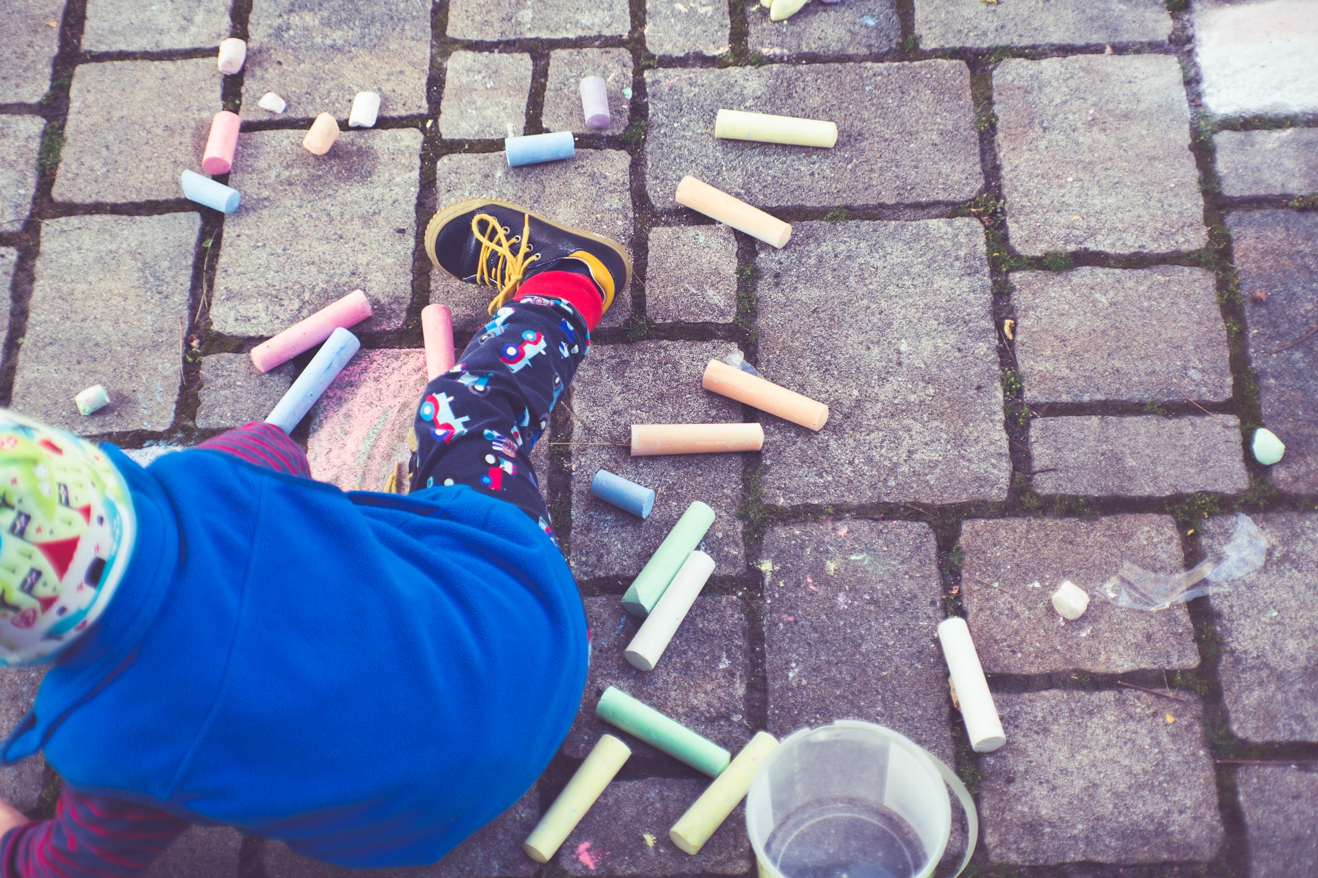Child with colorful chalk on brick patio, wearing blue sweater, patterned pants, and mismatched socks.