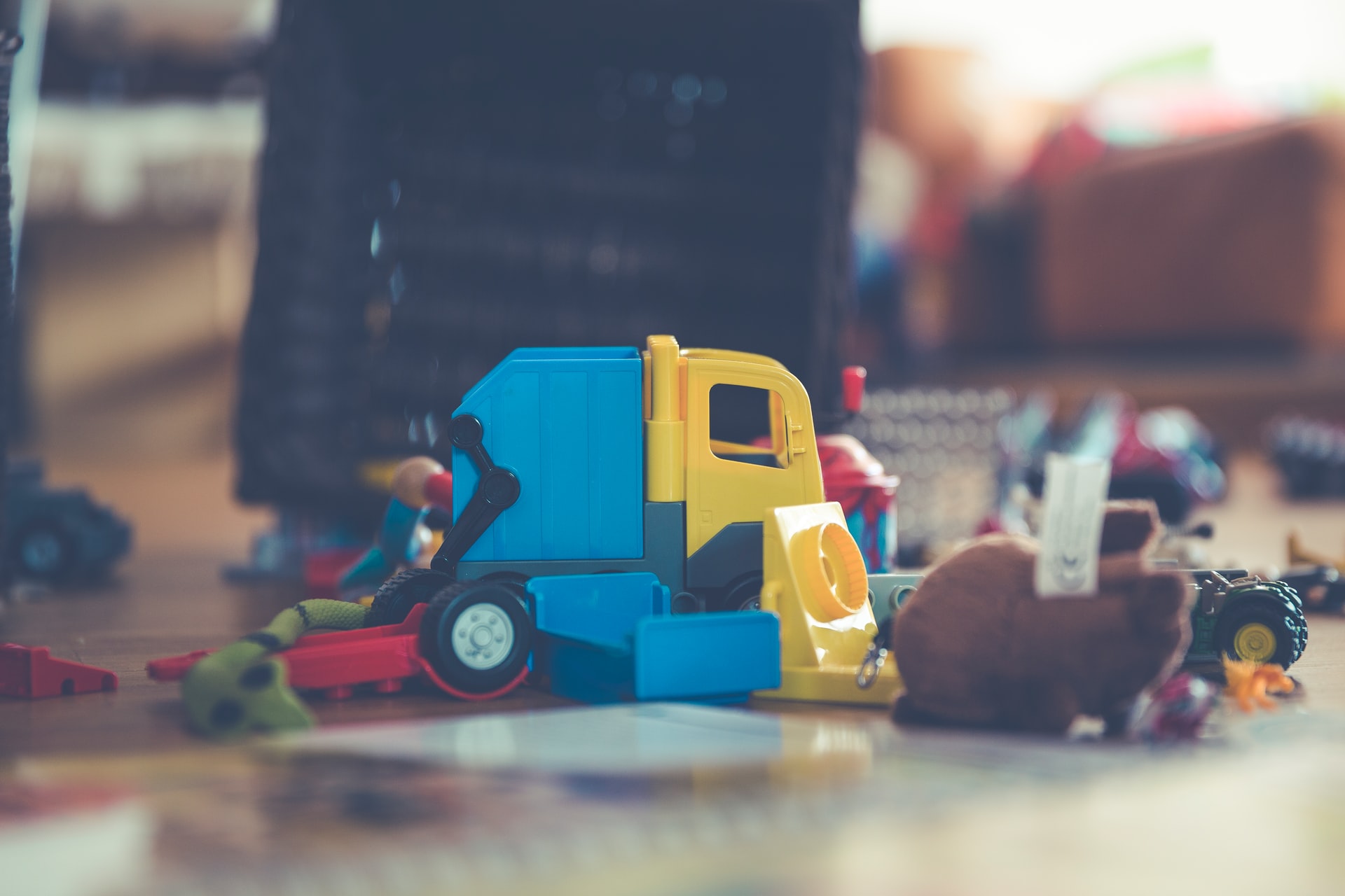 Toy truck, blue and yellow, on a wooden surface, surrounded by other toys in a blurred setting.