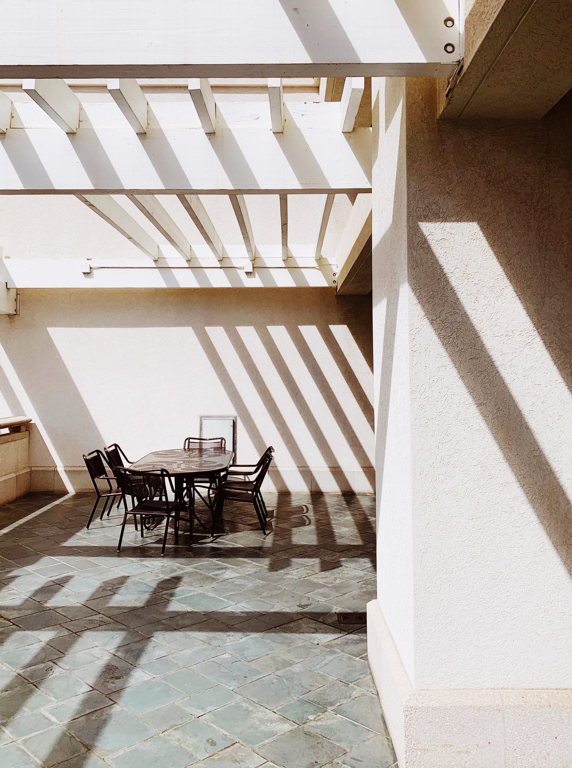 Patio with table and chairs under a white pergola, casting striped shadows on the floor and wall.