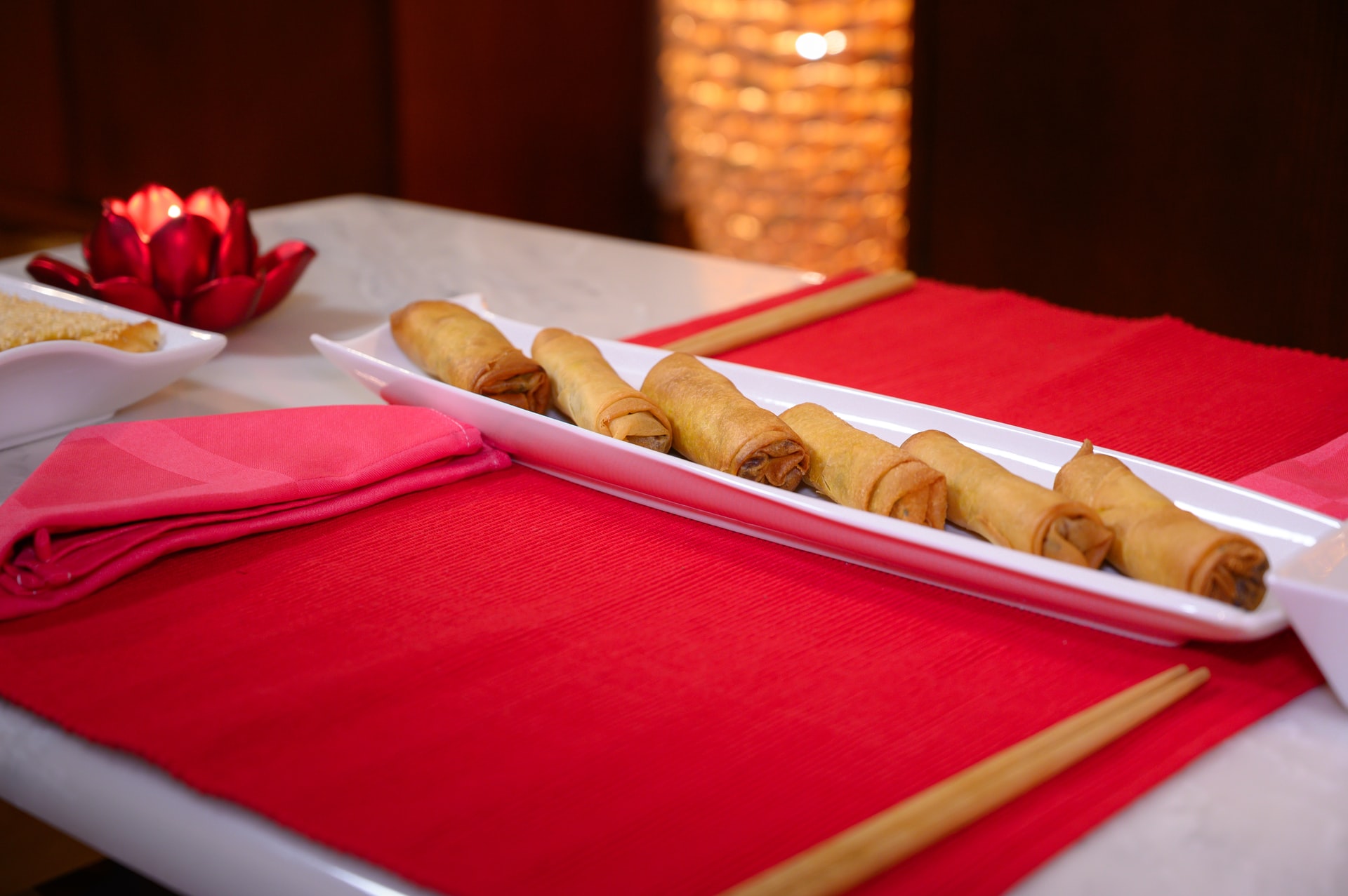 Spring rolls on a white platter on a red placemat with chopsticks; dimly lit setting.